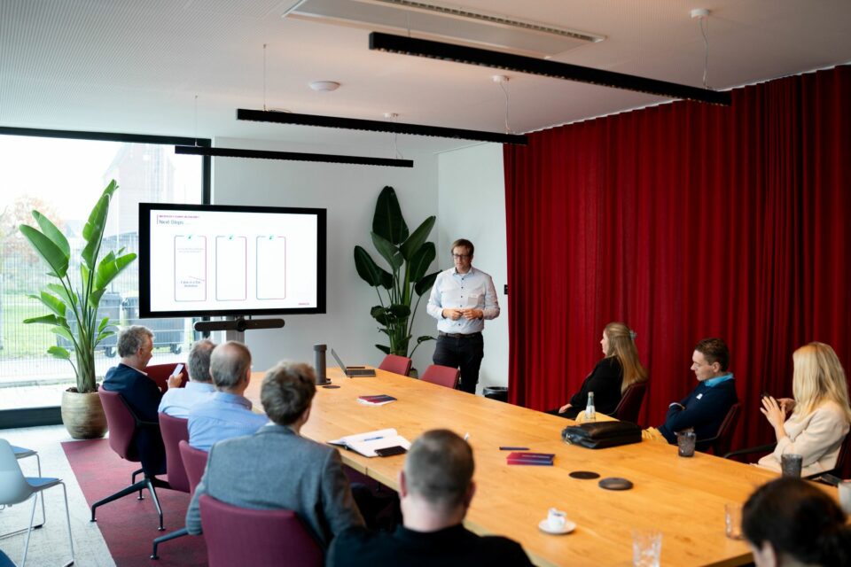 Data Driven Retail Day - Blick in einen Raum mit Personen, die an einem Konferenztisch sitzen dem Vortragenden zuhören.